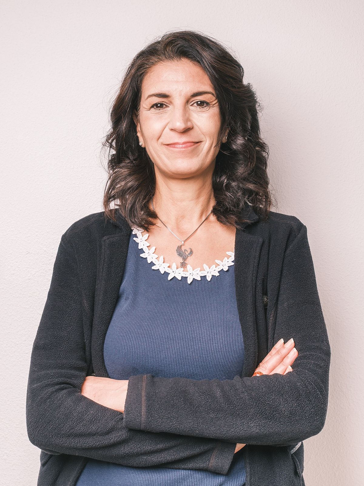 Femme aux cheveux bruns bouclés, les bras croisés, portant un blazer foncé sur un haut bleu, sur fond clair.