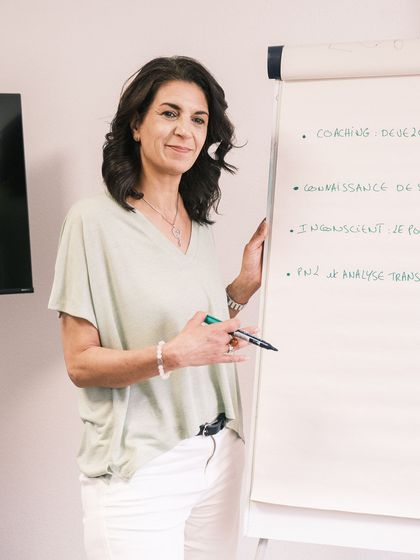 Femme en haut clair et pantalon blanc présentant un exposé sur un tableau de conférence avec un marqueur.