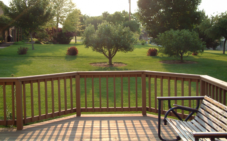 A photo of a deck that overlooks a beautiful green backyard. The deck has a railing installed to prevent people from falling off the side.