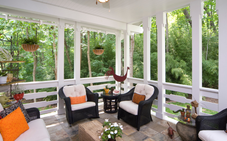 A photo of an enclosed porch in the backyard of a house. The patio has cozy chairs, a table and flowers. 