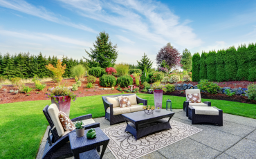 A beautiful photo of a luxurious patio in the backyard of a home. The patio has tables, chairs, and a rug sitting on it. In the distance beyond the patio there is a stunning flower bed with over 50 different types of flowers and plants