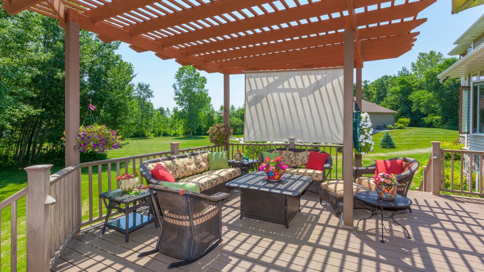 A picturesque wooden deck in San Bernardino, CA, is shaded by a charming pergola and adorned with a variety of comfortable seating options. Floral-patterned cushions and vibrant red and green throw pillows enhance the inviting atmosphere. A black outdoor table with colorful plant decorations serves as the focal point of the seating area. Surrounding the deck are lush green lawns and trees, creating a serene backdrop. Hanging baskets with blooming flowers and a rolling shade add both aesthetic and functional elements to this outdoor living space.