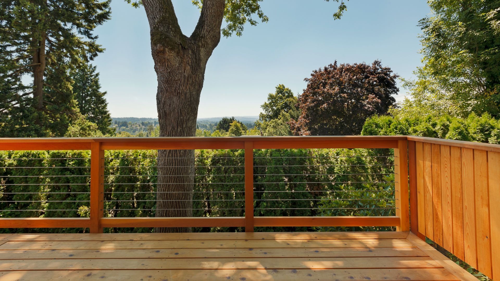 Scenic view from a wooden deck in San Bernardino, CA, overlooking a lush landscape filled with trees and greenery. The deck features simple wooden railings with horizontal metal cables, offering an unobstructed view of the natural surroundings. A large tree nearby adds to the serene atmosphere, providing shade and a connection to nature. The tranquil setting is perfect for outdoor relaxation and enjoying the picturesque scenery, showcasing the beauty and peacefulness of San Bernardino.