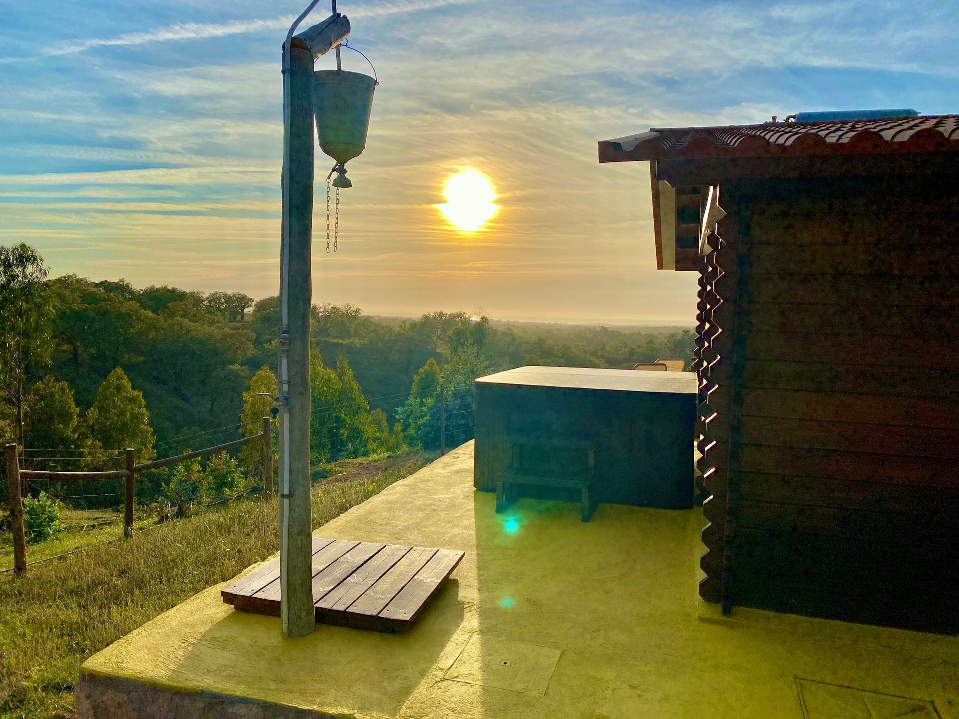 The sun is setting behind a wooden house with a hot tub in the background.