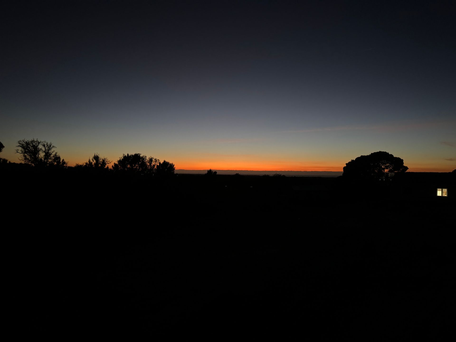A sunset over a field with trees in the foreground and a house in the background.