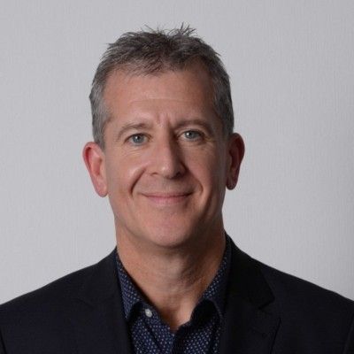 Man with gray hair, wearing a dark jacket over a patterned shirt, smiling.