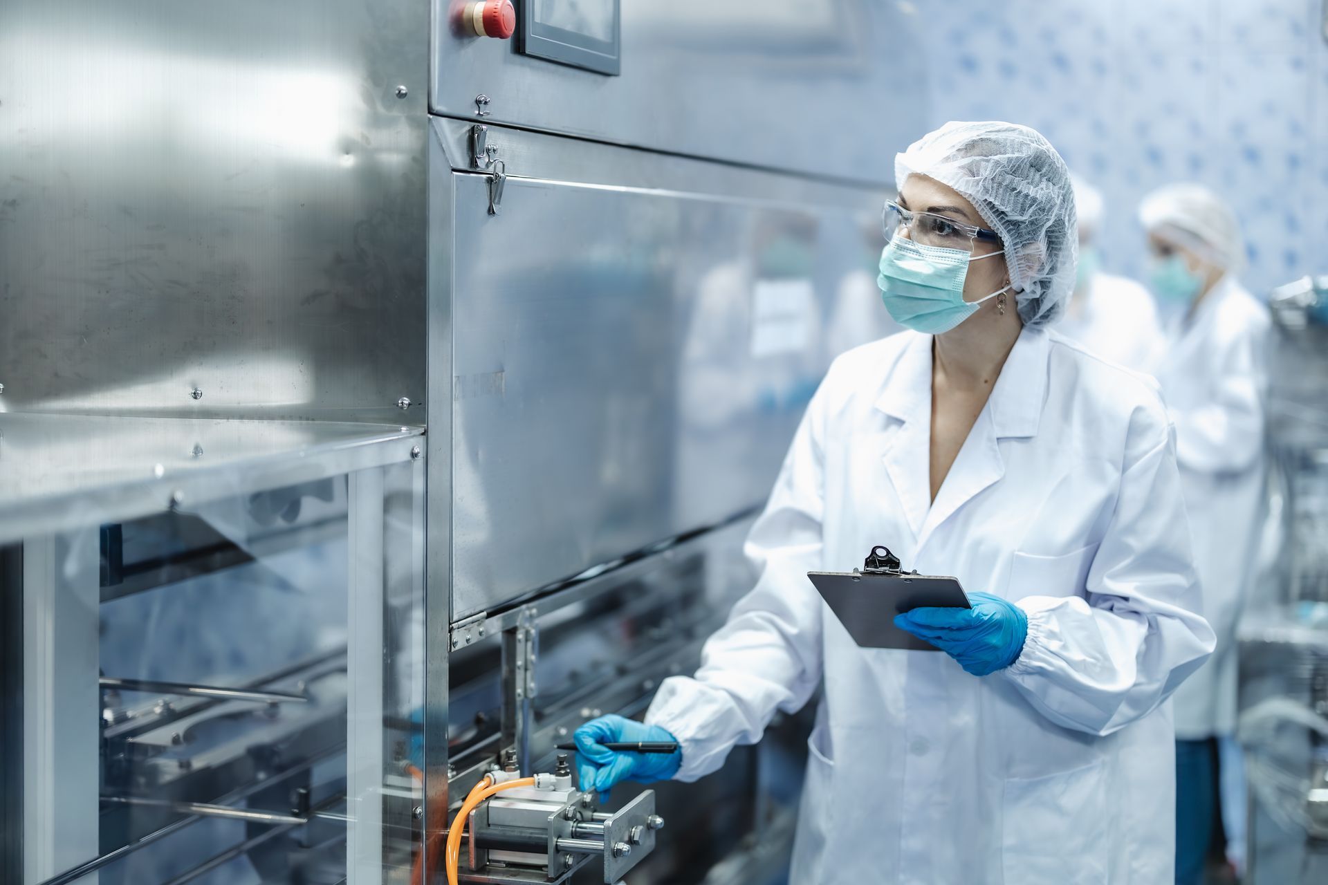 Woman in lab coat, mask, and gloves operates machinery in a factory. Other workers in background.