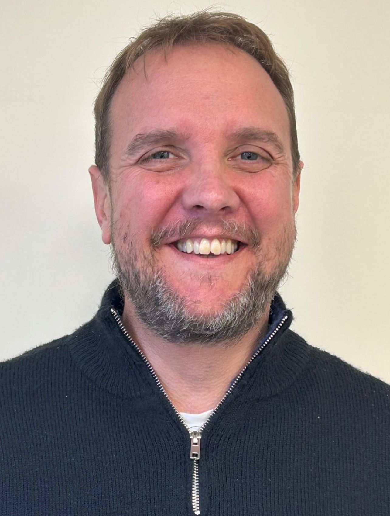 Man with a beard smiles at the camera, wearing a black zip-up sweater against a white background.