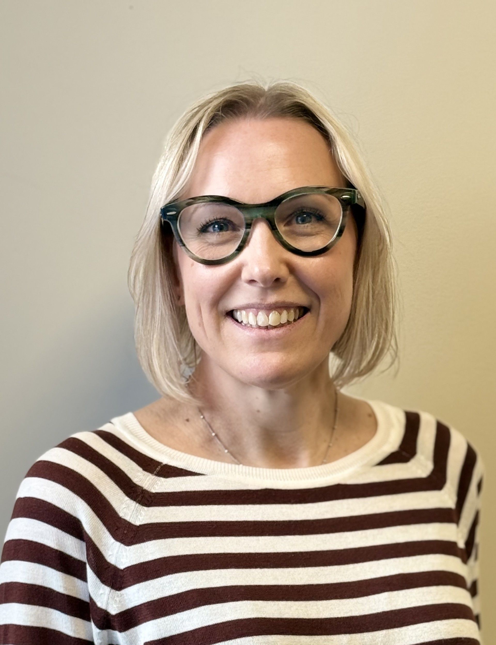 Woman with blonde hair and glasses smiles; wearing striped sweater, against a plain background.