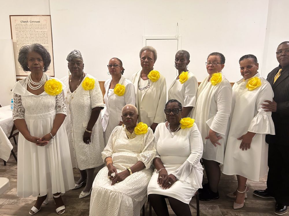 A group of women wearing white dresses and yellow flowers are posing for a picture.