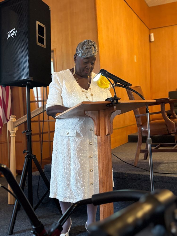 A woman in a white dress stands at a podium speaking into a microphone