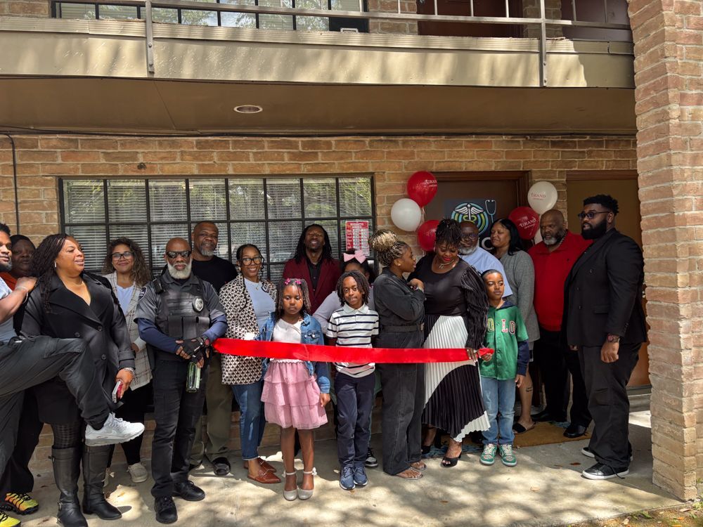 A large group of people are standing in front of a brick building holding a red ribbon.