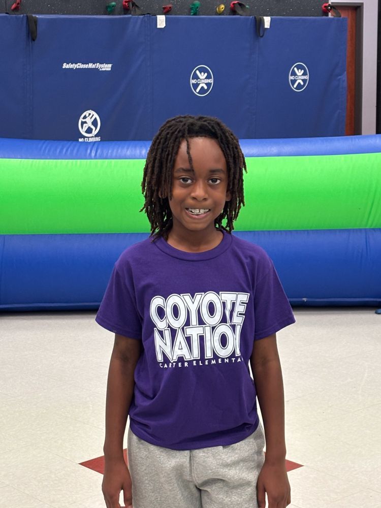 A young boy wearing a purple coyote nation shirt is standing in front of a bouncy house.