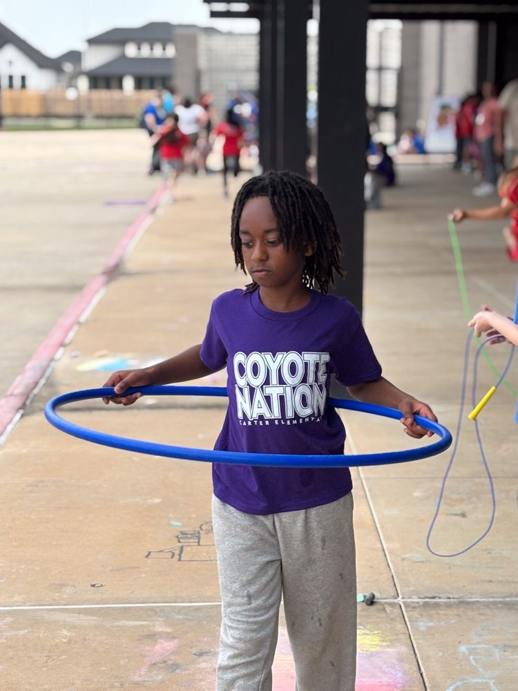 A young boy wearing a purple coyote nation shirt is holding a blue hula hoop.