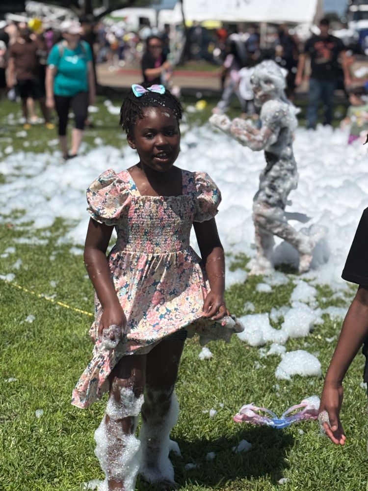 A little girl in a dress is standing in the grass covered in foam.