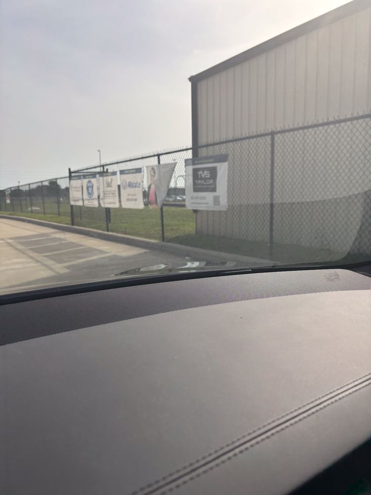 A car is driving down a road with a fence and a building in the background.