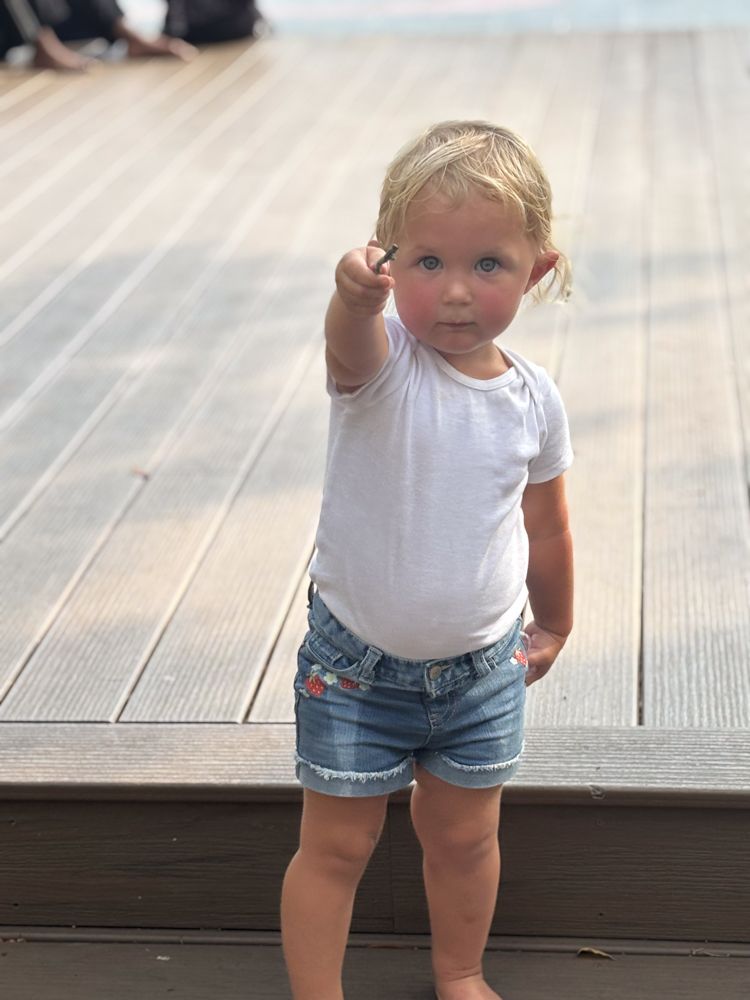 A little girl in a white shirt and denim shorts is standing on a wooden deck.