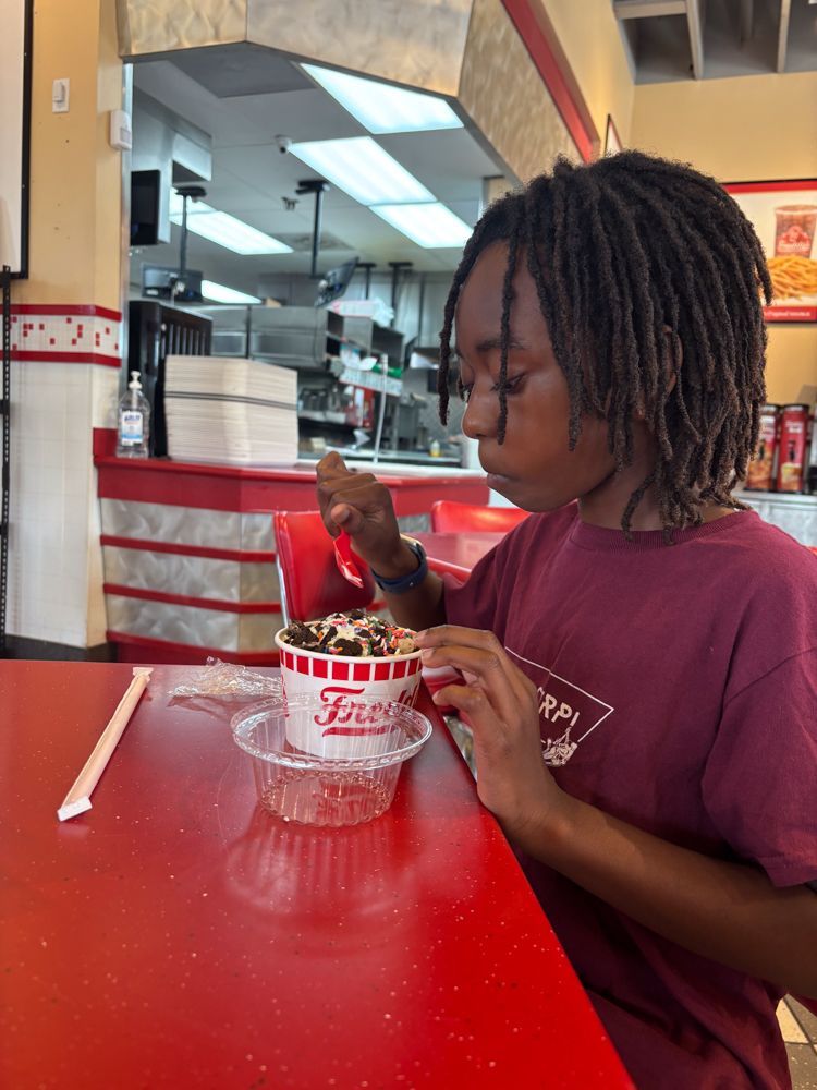 A young boy is sitting at a table eating ice cream from a fred 's cup.