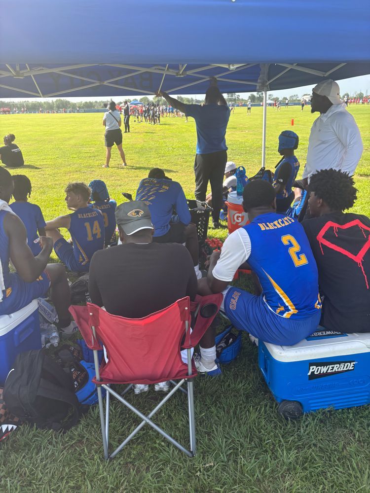 A group of people are sitting under a blue tent on a field.
