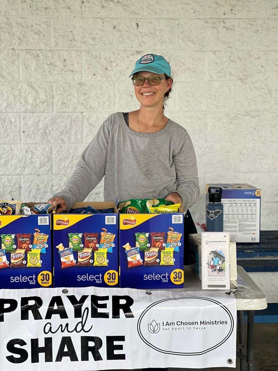 A woman is standing behind a table with boxes of chips and a sign that says prayer and share