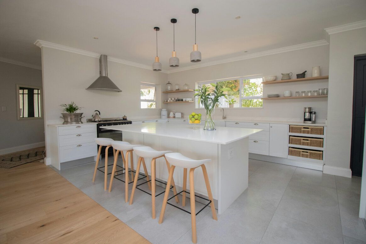 Modern white kitchen with island, stools, and pendant lights. Light wood floors, shelves, and accents.