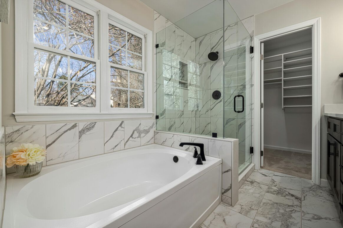 Modern bathroom with white tub, glass shower, and walk-in closet. Marble-look tile and black fixtures.