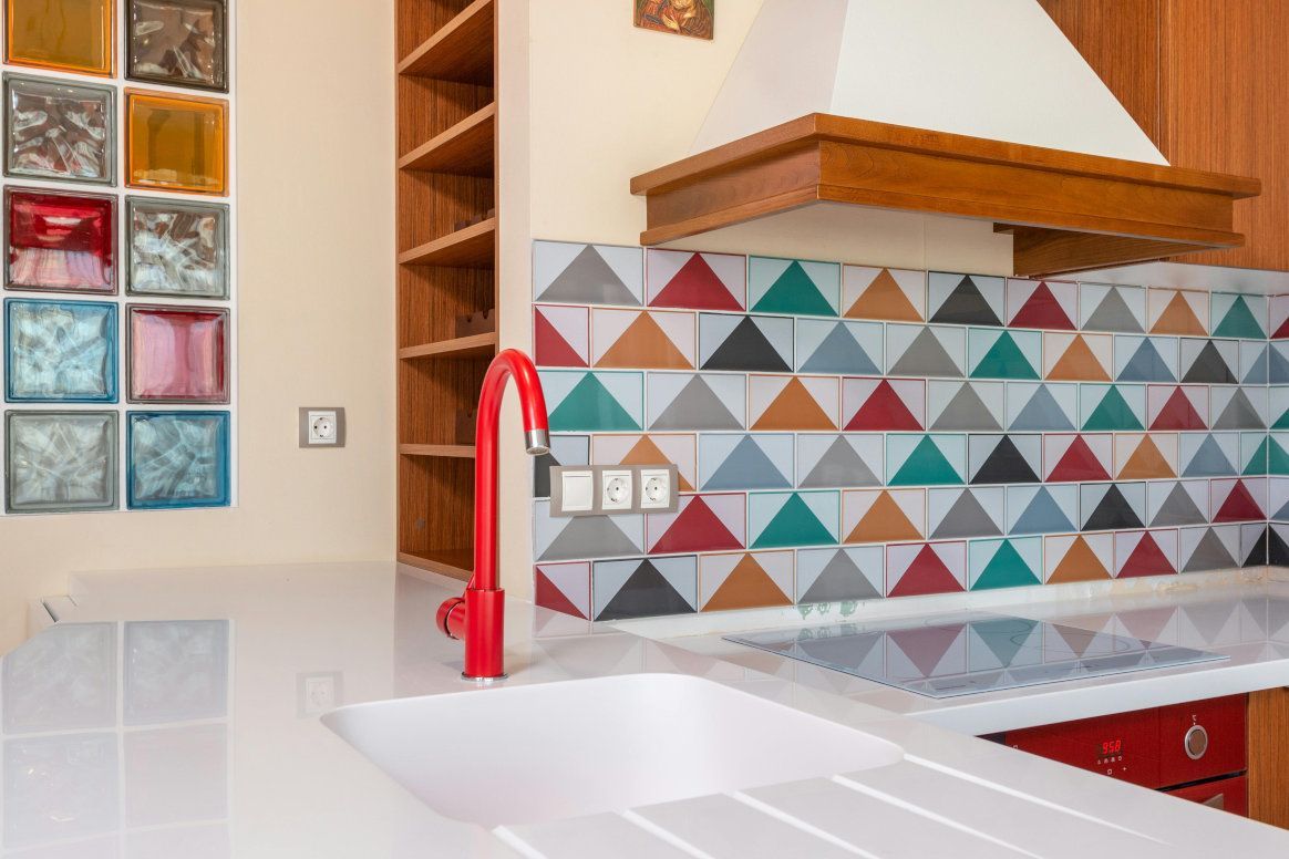 Kitchen with red faucet, geometric backsplash, white countertop, and wooden cabinets.