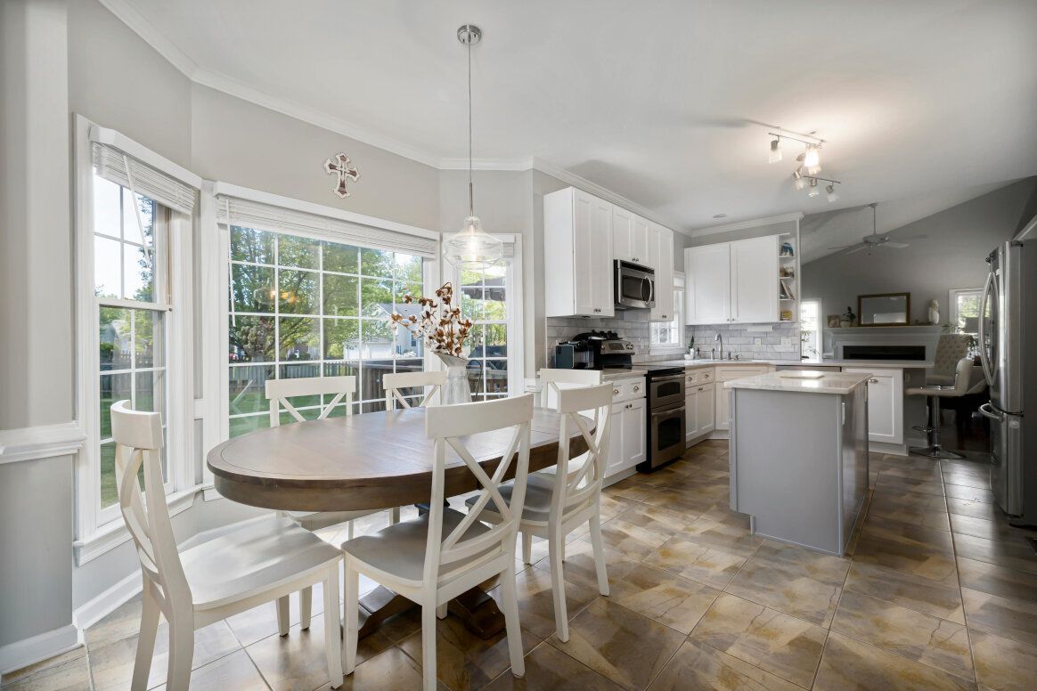 Kitchen with a round wooden table and white chairs by a large window. White cabinets, island, and stainless steel appliances.