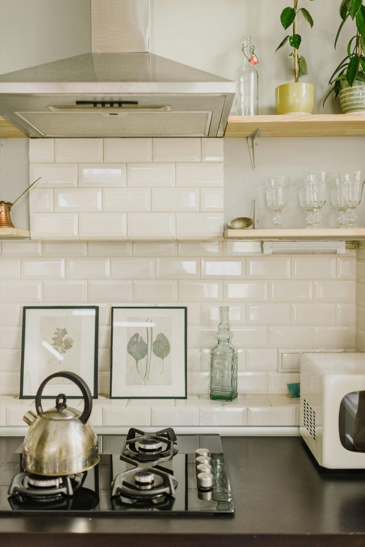 Kitchen with stainless steel range hood, black stovetop, white tile backsplash, open shelves, and framed botanical prints.