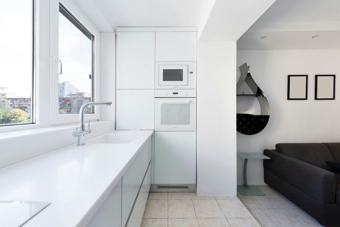 Bright white modern kitchen with built-in appliances, sink by window, and view into a living area.