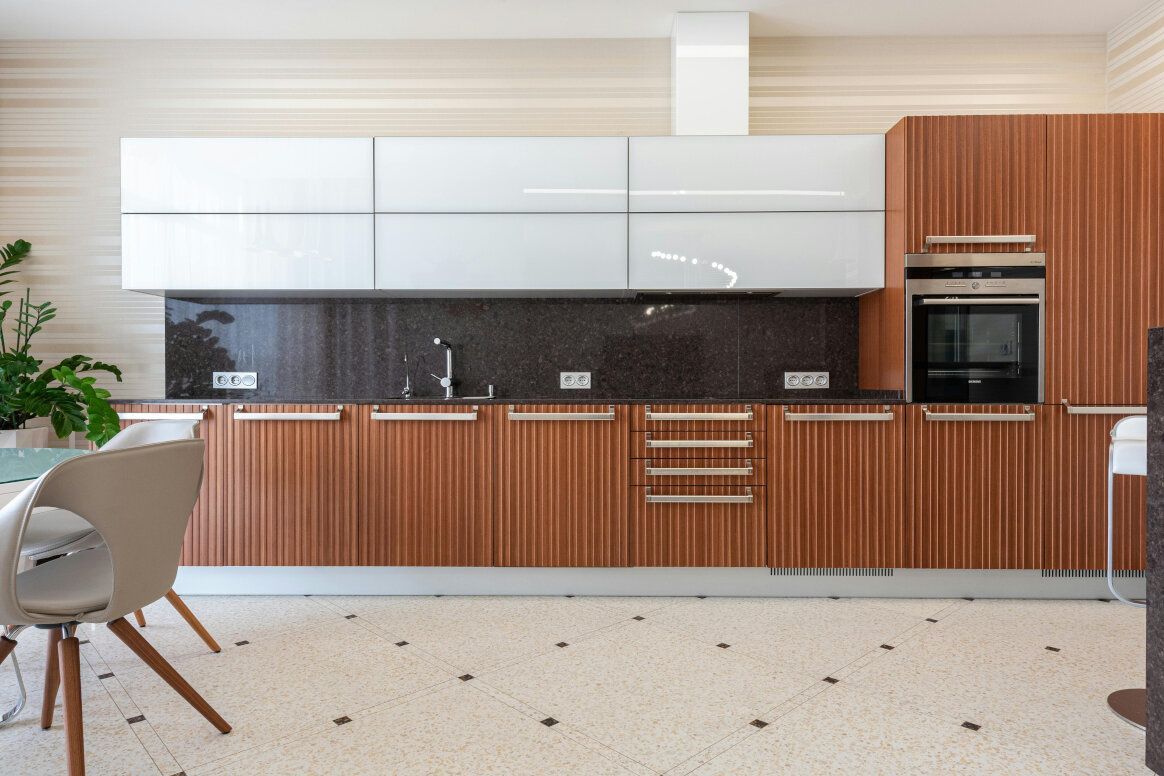 Modern kitchen with wood cabinets, white uppers, and speckled flooring. Built-in oven, and sink against a dark backsplash.