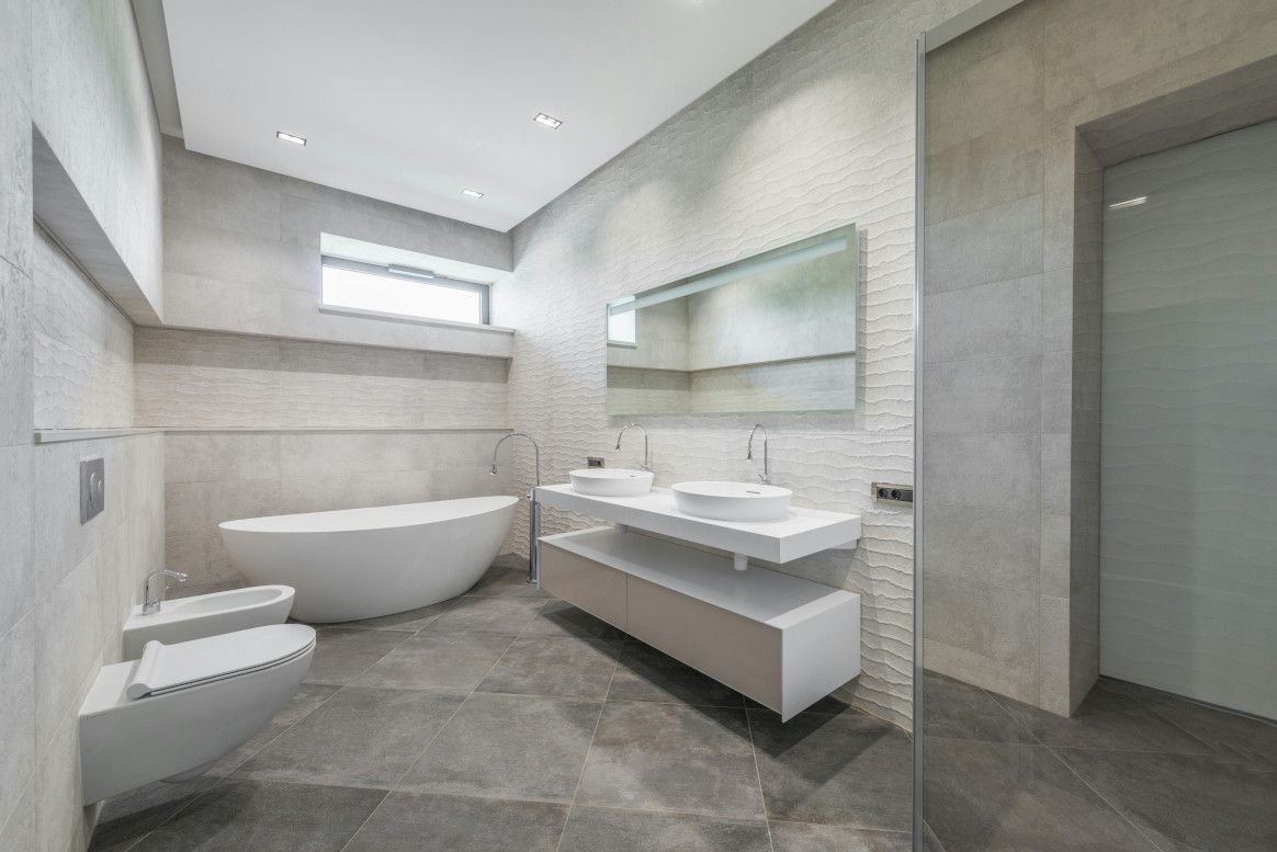 Modern bathroom with a bathtub, dual sinks, and toilet. Gray walls and floors, bright lighting.