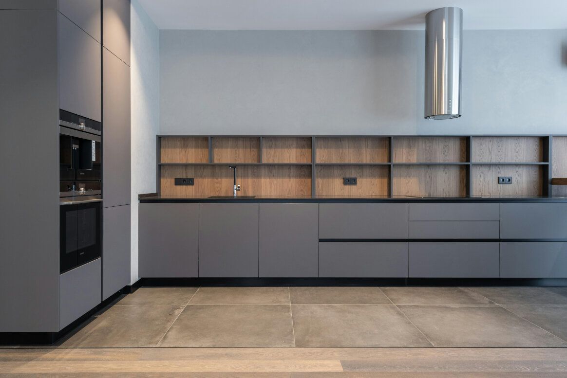 Modern kitchen with gray cabinetry, wood shelving, stainless steel range hood, and stone tile flooring.