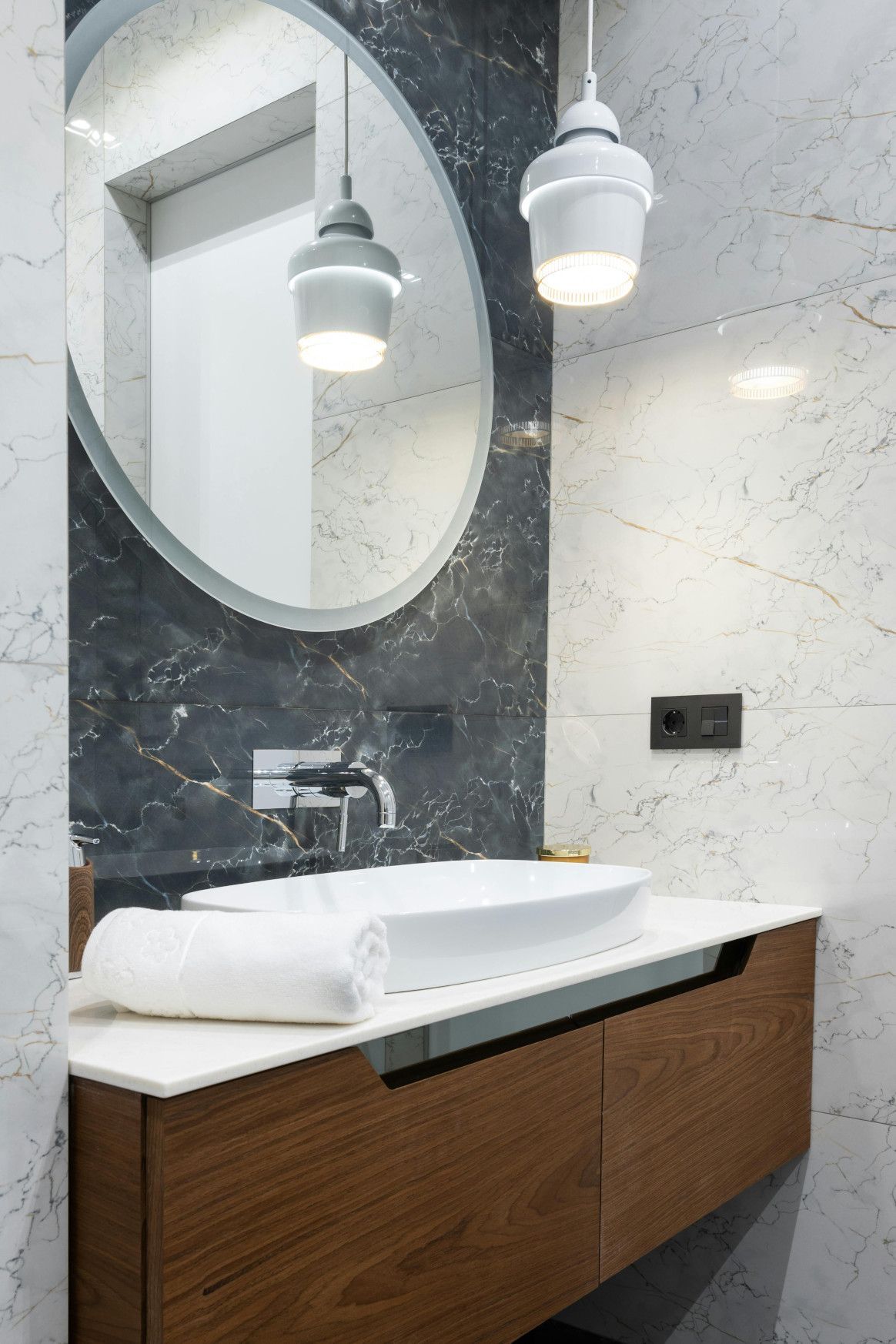 Modern bathroom with a white sink, wooden cabinet, and oval mirror. Two pendant lights hang overhead.