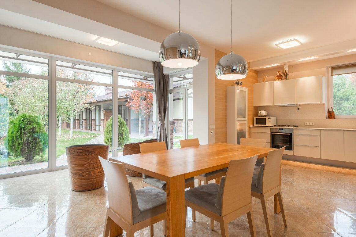 Bright dining area with wooden table, chairs, and a kitchen. Large windows and modern pendant lights.