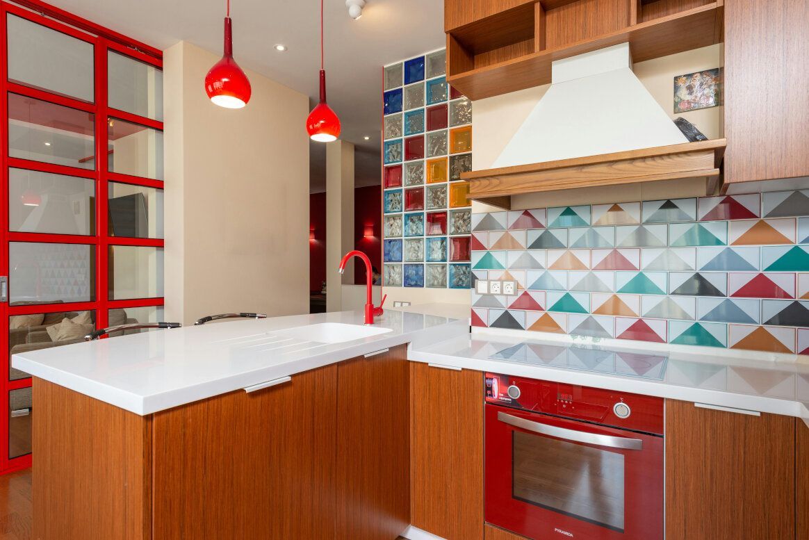 Modern kitchen with red accents; island, cabinets, oven. Colorful triangle and square tile backsplash.