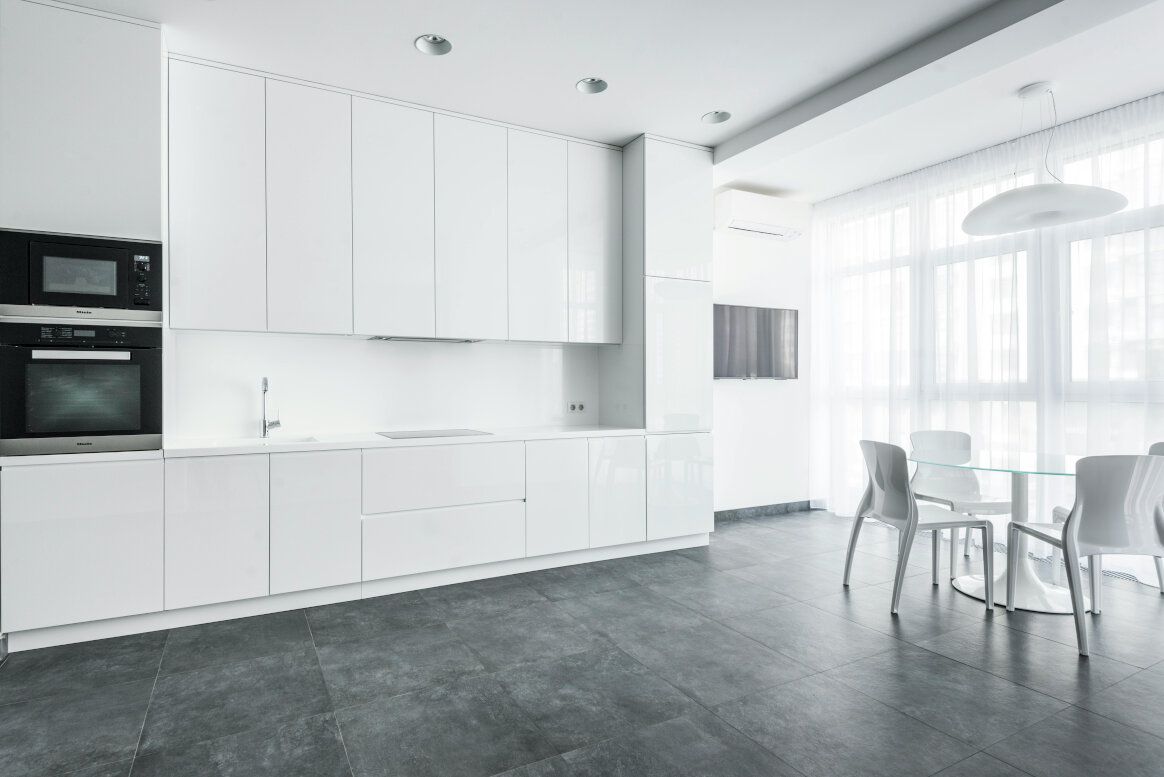 Modern all-white kitchen with cabinets, appliances, table, and chairs. Dark grey floor, bright window.