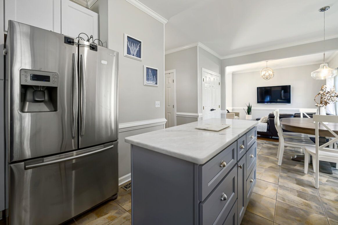 Stainless steel refrigerator, gray kitchen island with white countertop, dining area in the background.