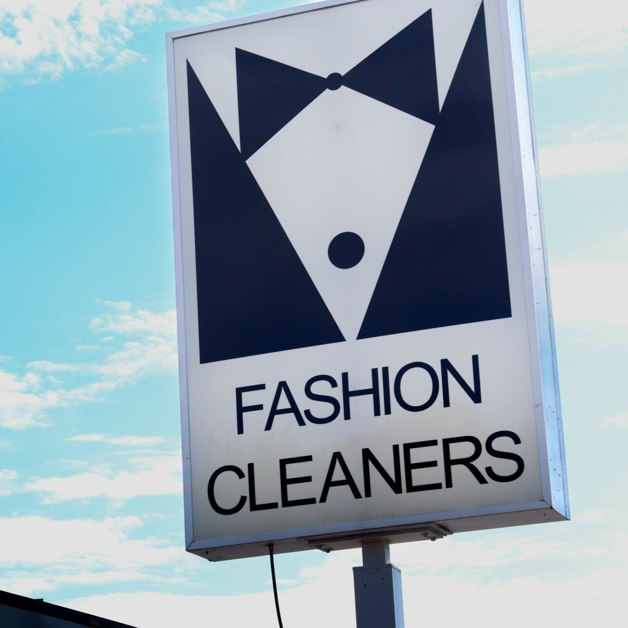 Sign for Fashion Cleaners with a black tuxedo graphic on a white background, against a blue sky.