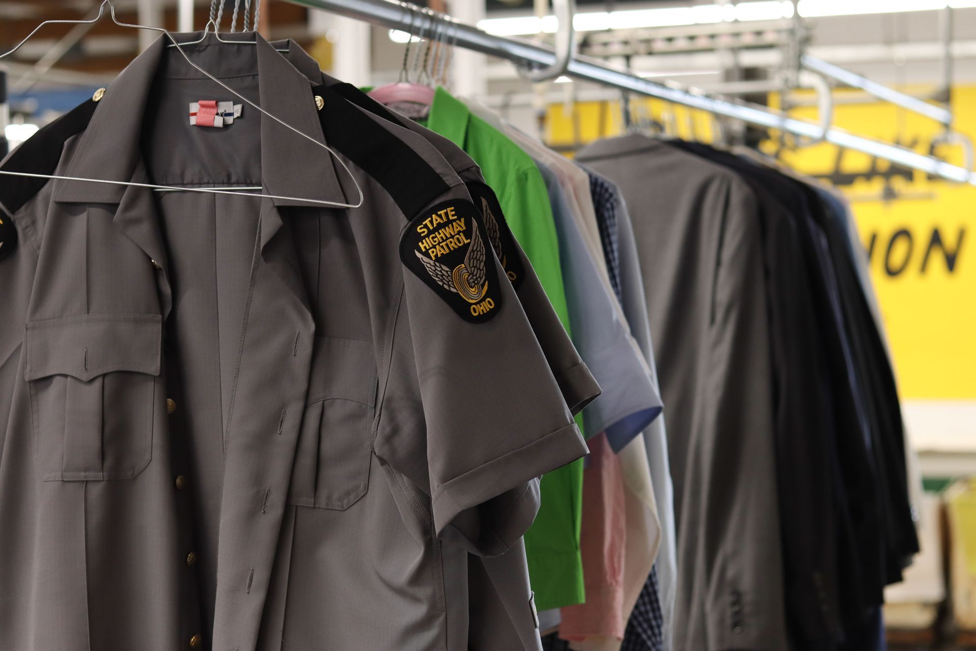 Clothes hanging on a rack, including a gray uniform with a patch, in a dry cleaning establishment.