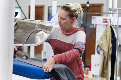 Woman ironing clothes in a dry cleaner. She has blonde hair in a bun and wears a red and white shirt.