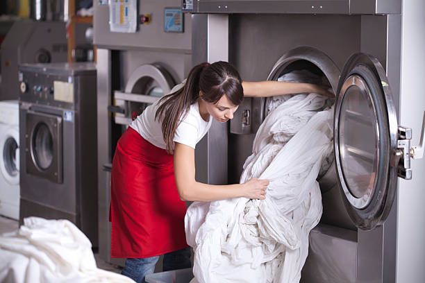 Woman in red apron loading laundry into a large commercial washing machine in a laundry facility.