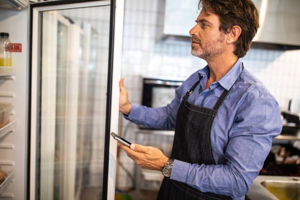 Man in apron checks refrigerator with phone in a kitchen.