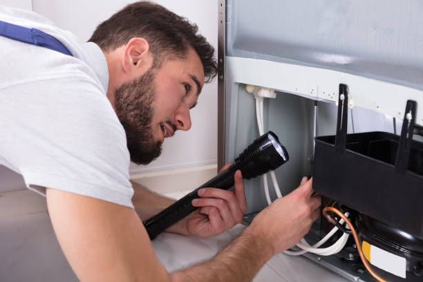 A repairman examines a refrigerator compressor with a flashlight.