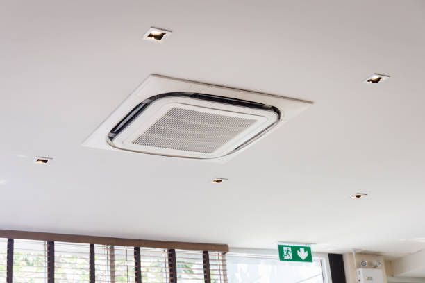 Ceiling-mounted air conditioner centered in a white ceiling with recessed lights and an exit sign.