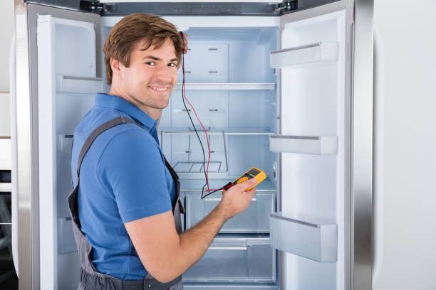 Man repairing refrigerator with a multimeter; smiling, wearing blue shirt and overalls.