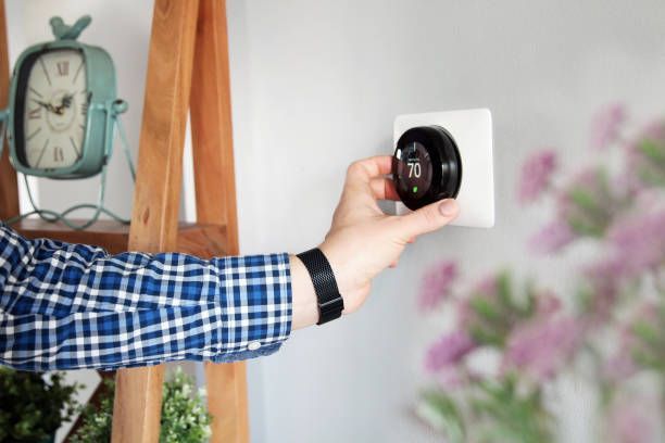 Hand adjusting a smart thermostat on a white wall, clock and flowers in the background.