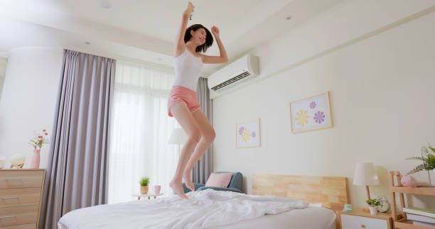 Woman jumping on a bed in a bright bedroom, wearing headphones, shorts and a tank top.