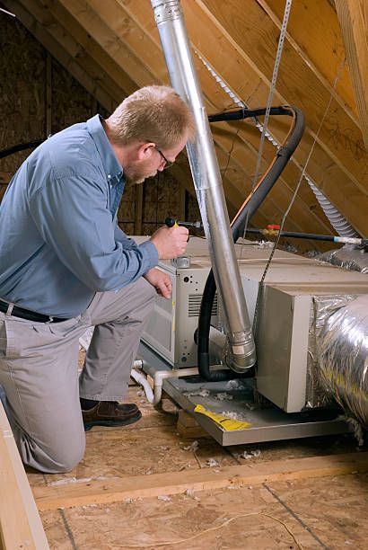 A person kneels, inspecting HVAC unit in an attic. They are holding a flashlight.