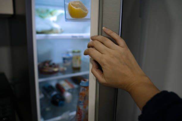 Hand opening refrigerator door; visible items inside lit by interior light.